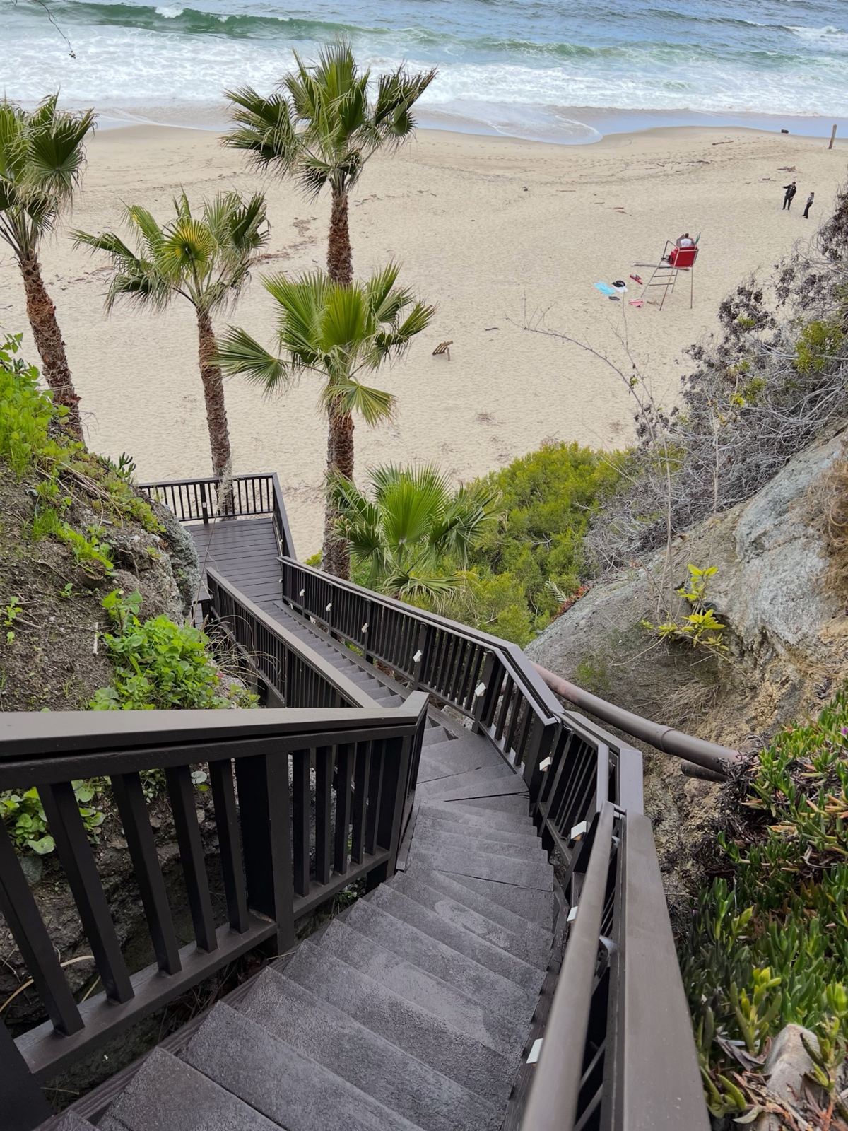 Dark brown exterior staircase and deck landings descending a bluff to the beach, with palm trees and the ocean below