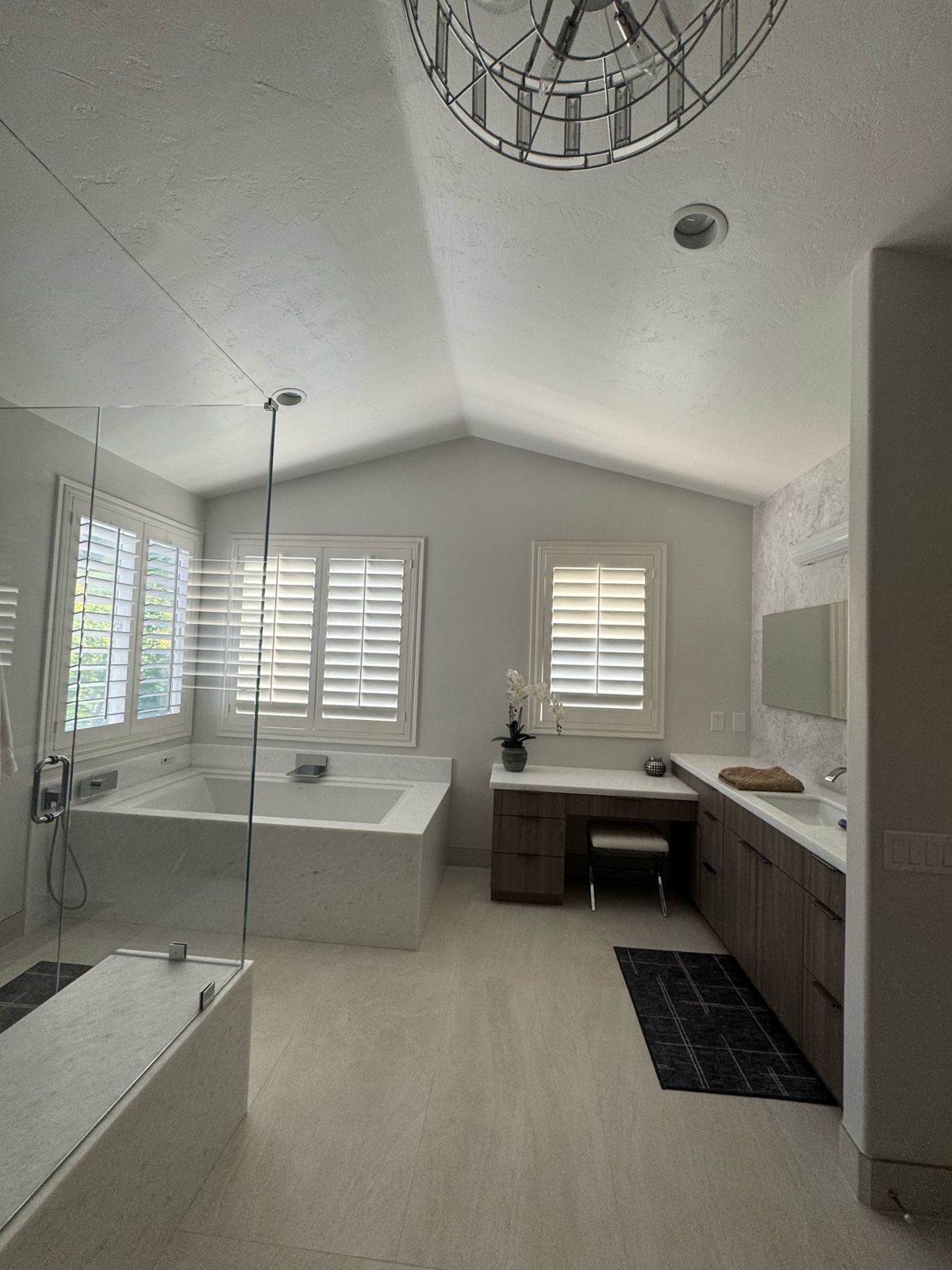Master bathroom with a vaulted textured ceiling, soft gray painted walls, plantation shutters, soaking tub, and wood vanity cabinets