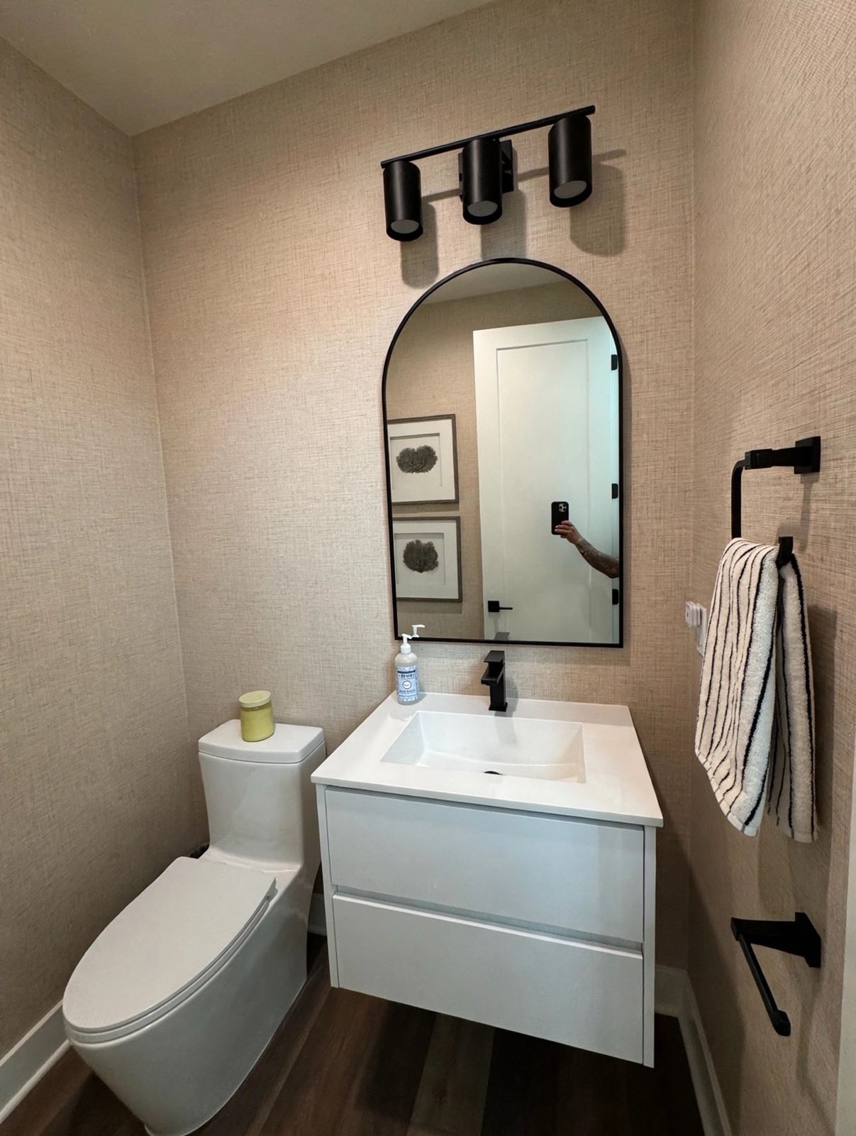 Powder room with beige grasscloth wallcovering, arched black-framed mirror, floating white vanity cabinet, and black fixtures