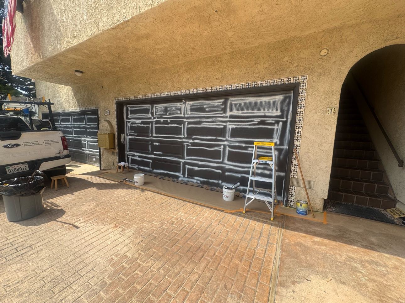 Two garage doors in the sanding and prep stage with white sanding marks, buckets, and a ladder on the driveway