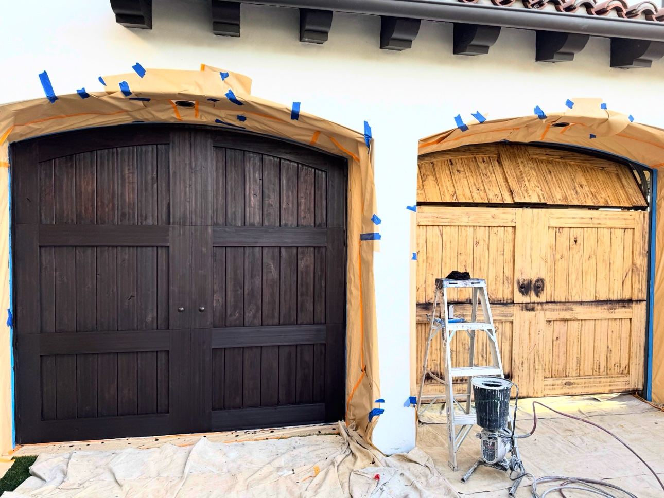 Two wood garage doors masked with paper and tape for staining, the left door finished dark and the right still raw wood with a sprayer and ladder in front