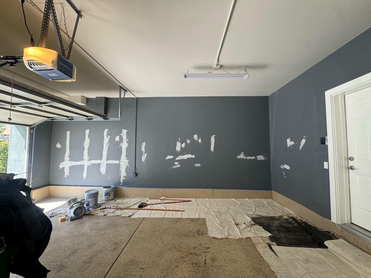 Garage walls with white drywall patch repairs over the slate-blue paint, drop cloths down, during prep