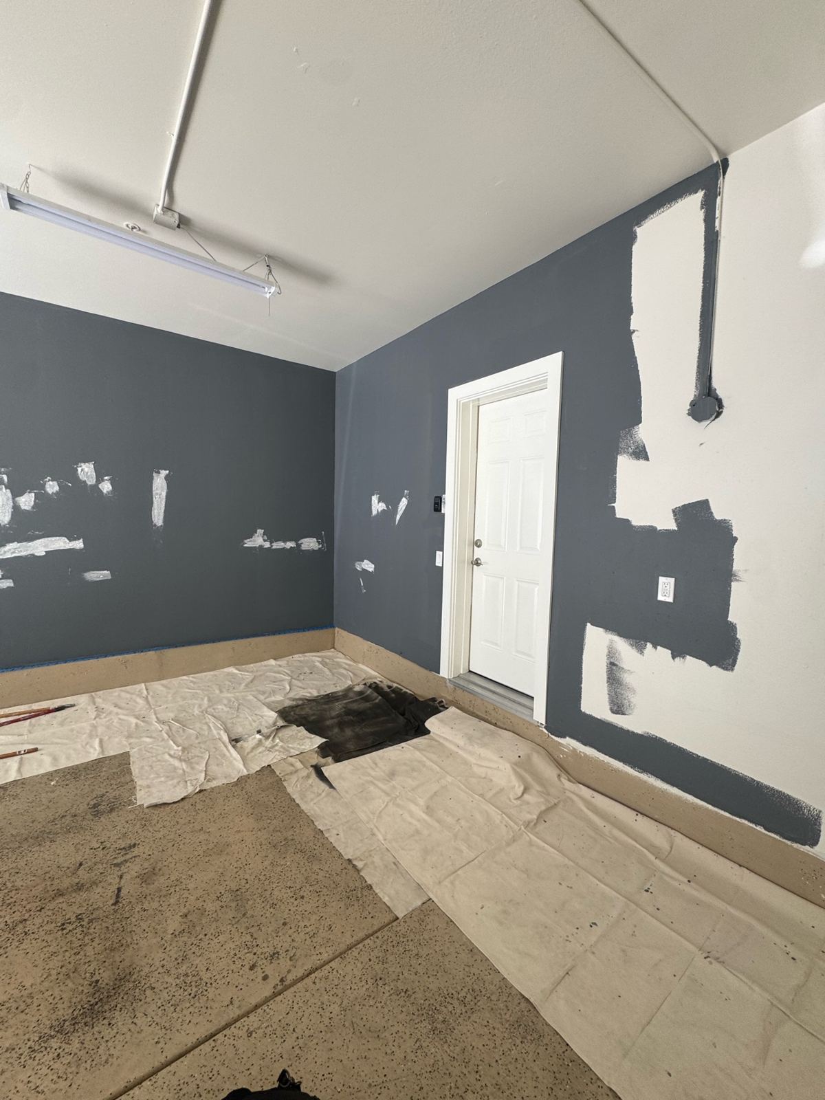 Garage wall being cut in with slate-blue paint around a white door, with patched spots and drop cloths down
